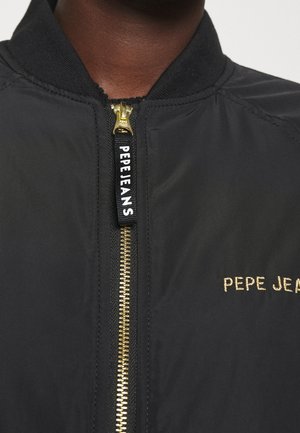 Czarna kurtka z złotym zamkiem błyskawicznym i metką "PEPE JEANS" na suwaku oraz wyszywanym logo na piersi, noszona przez osobę o ciemnej karnacji.