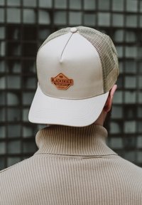 Gorra trucker beige con parte trasera de malla y un parche de cuero marrón cosido que dice "BLACKSKIES". Cuello de suéter ribeteado en color beige al fondo.