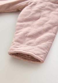 Maniche in tessuto trapuntato di un delicato rosa, caratterizzate da una superficie testurizzata con cuciture sottili e un polsino arrotondato. Vista ravvicinata che mette in risalto il materiale e il design.