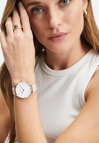 Roségoldene Uhr mit weißem Zifferblatt und minimalistischen Stundenmarkierungen, ausgestattet mit einem hellbeigen Armband mit geprägten Details; am Handgelenk getragen.