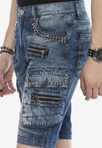 Hombre que lleva pantalones cortos de mezclilla azul con múltiples bolsillos con cremalleras y tachuelas, pulsera de cuentas negras en la muñeca izquierda y reloj negro en la muñeca derecha.