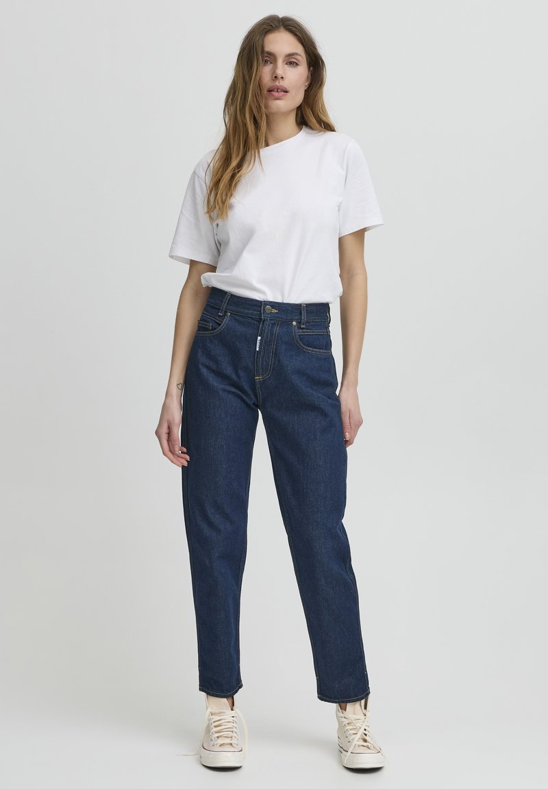 Jean taille haute bleu foncé avec une coupe droite, quatre poches, fermeture à bouton en métal argenté et des détails de couture discrets, associé à un t-shirt blanc.