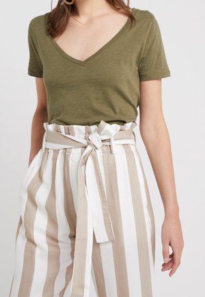 Femme portant un haut vert olive à manches courtes et col en V, rentré dans un pantalon taille haute blanc et beige à rayures avec une ceinture en tissu nouée.