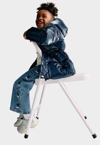 Fodrad marinblå puffjacka, vit topp och jeans med fjärilsmönster. Barn sitter på en vit hopfällbar stol, grå sneakers synliga.