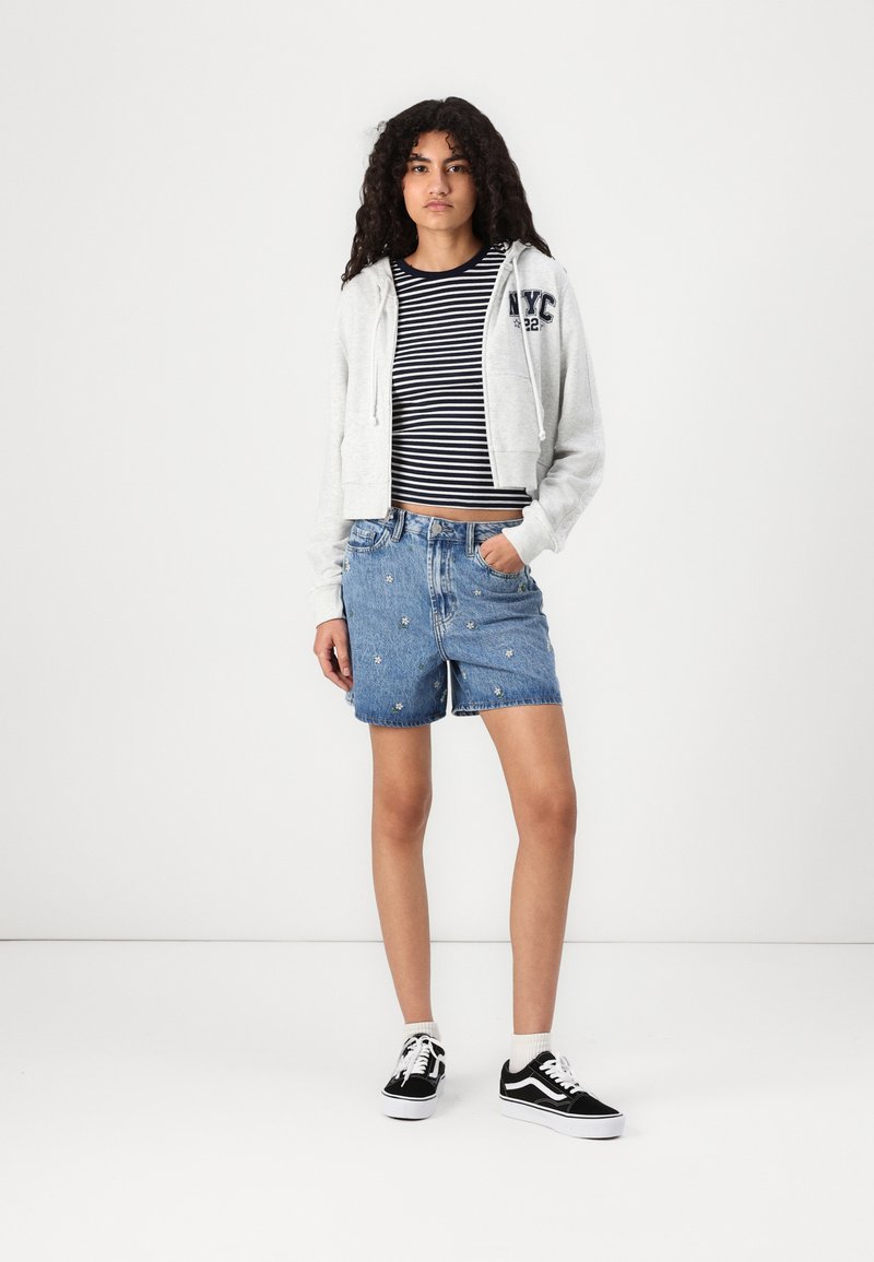 Felpa grigia corta con cappuccio e lacci, camicia a righe blu e bianche, pantaloni shorts di jeans ricamati e sneakers nere con suola bianca.