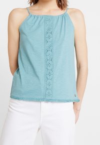 Haut sans manches bleu clair avec un panneau en dentelle au centre et un ourlet à franges. Confectionné dans un tissu doux, doté de fines bretelles.