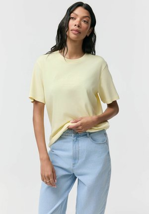 Kobieta z długimi, ciemnymi włosami nosi jasnobeżową koszulkę z krótkim rękawem i jasnoniebieskie dżinsy z wysokim stanem, trzymając brzeg koszulki jedną ręką.