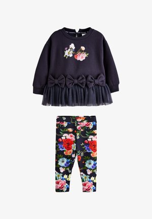 Unge barn står iført en sort blomstret sweatshirt med sløjfer og tyl, blomstrede leggings og hvide velcro-sneakers på en ensfarvet baggrund.