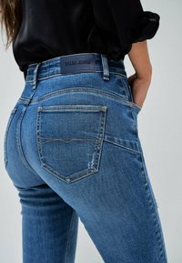 Jeans de mezclilla azul ajustados con cintura de altura media, que presentan un parche de logo de cuero y bolsillos traseros clásicos con acentos de costura curvada.