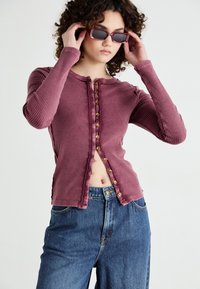 Ribbed long-sleeve top in dusty rose with a round neckline, button front detail, and frayed hem, paired with high-waisted blue jeans.