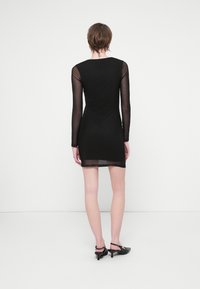 Rochie neagră cu mâneci lungi din material transparent, design ajustat, lungime până la mijlocul coapsei, textură netedă și spate simplu. Poartă pantofi cu toc negri.