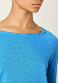 Mėlyna ilgomis rankovėmis palaidinė su plačiu iškirpimu ir tekstūruota audiniu. Savybės - nebaigtas kraštas iškirpimo ir peties siūlėse.