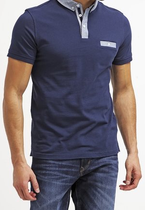 Man draagt een marineblauw poloshirt met korte mouwen en een contrasterende lichtblauwe kraag en zakklep, gecombineerd met donkerblauwe jeans.
