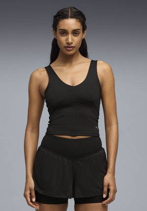 Γυναίκα με πλεξούδες, που φοράει μαύρο αθλητικό τοπ χωρίς μανίκια και μαύρο σορτς πολλαπλών στρωμάτων, στο φόντο γκρι διαβαθμισμένου χρώματος.