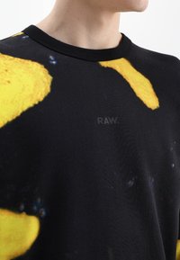 Sudadera negra con un gran patrón abstracto amarillo, cuello redondo y un pequeño logo "RAW." impreso en negro en la parte frontal.