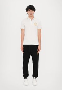 Balta polo marškinėliai su geltonais akcentais ir logotipu, derinami su juodomis sportinėmis kelnėmis, puoštais auksiniu emblemą, užbaigti baltais sportiniais bateliais.