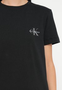 Melnā kokvilnas T-krekls ar apaļu kakla izgriezumu un īsām piedurknēm, kreisajā krūtīs ar sudraba Calvin Klein logotipu un monogrammu.