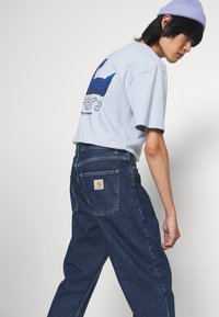 Oseba v svetlo modri majici z grafiko, temno modrih Carhartt kavbojkah in sivem klobuku, stoji ob ozadju brez posebnosti.