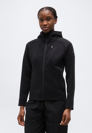 Kobieta z warkoczem, ubrana w czarną bluzę z zamkiem i czarne spodnie, stojąca na tle jednolitego jasnego tła.