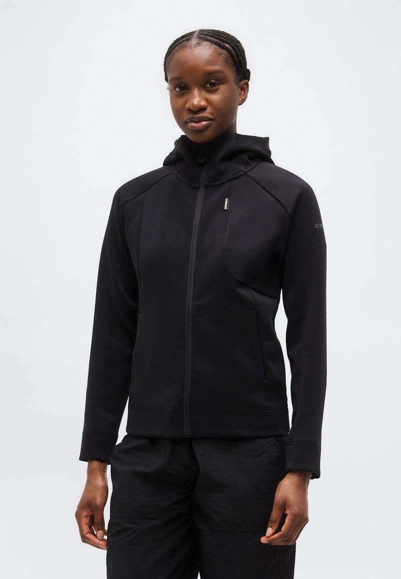 Γυναίκα με πλεξουδωτά μαλλιά, φοράει μαύρο φούτερ με φερμουάρ και μαύρο παντελόνι, στέκεται μπροστά σε ένα απλό φωτεινό φόντο.