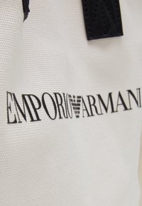 Tissu blanc texturé avec logo Emporio Armani noir et emblème d'aigle, présentant des détails de sangle en cuir noir cousus.