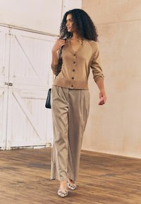 Cardigan beige avec fermeture à boutons, pantalons plissés beige clair et ballerines à imprimé serpent. Sac à main noir, cheveux texturés, fond en parquet.