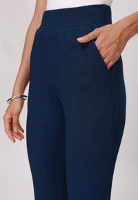 Navy ribbestrikkede leggings med høy midje, med sidelommer. Stoffet har en jevn tekstur og et konsekvent vertikalt mønster.