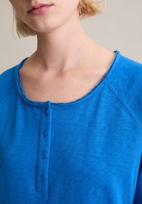 Niebieska koszula henley z zaokrąglonym dekoltem, widocznymi przeszyciami i czterema guzikami przy listwie. Miękka, lekko teksturowana tkanina.