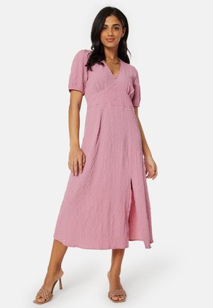 Abito midi rosa texturizzato con maniche a sbuffo, dettagli a bottoni e uno spacco davanti. Tessuto liscio con un leggero motivo a increspature.