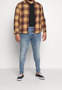 Hombre vistiendo jeans ajustados de color azul claro, zapatillas negras, camiseta negra y una camisa de cuadros amarilla y marrón abierta, de pie contra un fondo blanco.