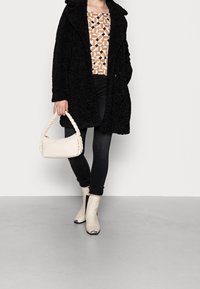 Manteau noir texturé, haut à motifs beige et marron, jean skinny noir, sac à main tressé blanc et bottines crème avec des accents noirs.