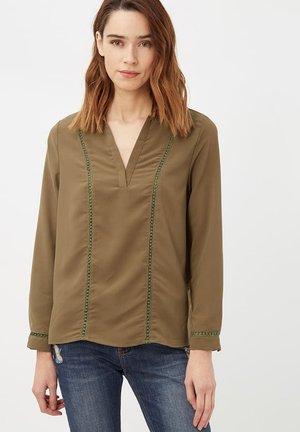 Mujer con cabello castaño hasta los hombros que lleva una blusa de manga larga verde oliva con costuras verticales decorativas y vaqueros azules.