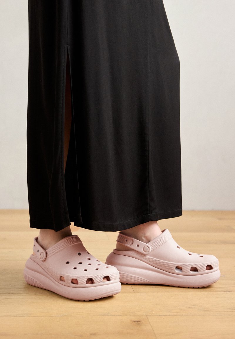 Zuecos de color rosa claro con agujeros de ventilación, suela gruesa y correa trasera, combinados con una falda larga negra y un fondo de suelo de madera.