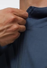 Veste bleu foncé avec un col haut et une fermeture éclair. Une main saisit la tirette de la fermeture éclair, mettant en valeur la texture lisse du tissu de la veste.