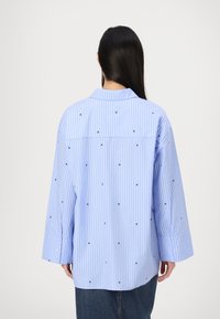 Chemise rayée bleue et blanche avec de grands poignets, ornée de petits détails carrés noirs dispersés. Tissu doux, coupe ample et empiècement au dos.