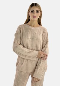 Cable-knit sweater in light pink, featuring a wide neckline and ribbed cuffs. The textured fabric has a soft feel, offering a relaxed fit.