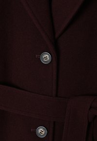 Bordeaux vlněný kabát s klopovým límcem, dvěma knoflíky a páskem v pase. Texturovaná tkanina s jemným povrchem.