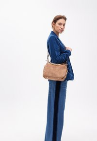 Una donna sta in posizione laterale, indossando un blazer blu e jeans a vita alta, mentre tiene una borsa beige con una trama in velluto a coste.
