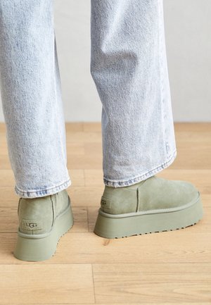 Person trägt hellblaue Jeans und olivgrüne UGG-Plateaustiefel, steht auf einem hellen Holzboden.