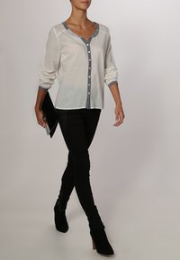 Camicia bianca con colletto a V profondo e rifiniture grigie, indossata con jeans skinny neri e stivaletti neri con tacco.