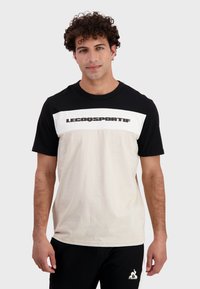 T-shirt da uomo con una sezione superiore nera, una sezione inferiore beige e una fascia orizzontale bianca con la scritta "LECOQSPORTIF" in grassetto nero.