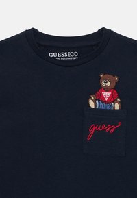 Námornícky modré tričko z organickej bavlny s vyšitým medveďom v červenom tričku na ľavej náprsnickej vrecku a červeným vyšitým textom "guess".