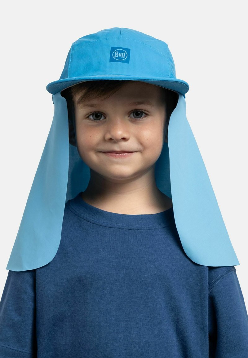 Niño pequeño con un sombrero azul para el sol con ala para el cuello y una camisa azul a juego, sonriendo suavemente sobre un fondo liso.