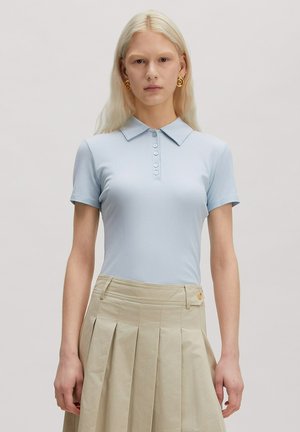 Blond vrouw met gouden spiraalvormige oorbellen, een lichtblauw overhemd met korte mouwen, kraag en knopen, en een beige geplooide rok.