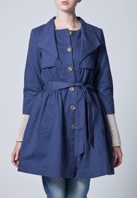 Granatowy płaszcz trencz z dużym kołnierzem, zapięciem na guziki, paskiem w talii i bocznymi kieszeniami. Posiada teksturowane akcenty z dzianiny na rękawach. Wykonany z bawełny.