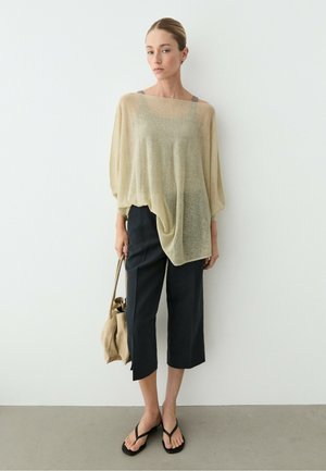Kvinde iført en beige løsstrikket top, sorte bukser i kort længde, sorte flip-flops, holder en beige taske, stående mod en helt hvid væg.