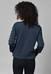 Sudadera azul marino de manga larga, cuello redondo y detalle gráfico blanco en la manga. Hecha de un tejido suave y texturizado. Combinada con pantalones negros.