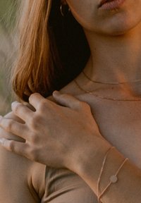 Roségoldene Lagenhalsketten und ein Armband mit einem kreisförmigen Charm, die elegant über eine kahle Schulter fallen, vor einem weichen, gedämpften Hintergrund.