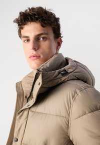 Veste rembourrée beige avec un col haut, une capuche amovible et une fermeture éclair apparente. Elle possède des panneaux cousus et des boutons-pression à l'avant.