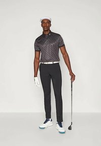 Μαύρο πουκάμισο polo με σχέδιο, μαύρο παντελόνι, λευκή ζώνη και λευκά παπούτσια γκολφ με μπλε λεπτομέρειες; κρατώντας ένα μπαστούνι γκολφ, φορώντας ένα λευκό καπέλο.